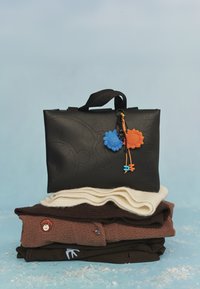 Zwarte matte tas met reliëfontwerp, bovenop kleurrijke gehaakte bedels. Eronder een stapel zachte truien in bruine en crèmekleuren.