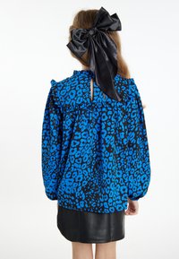 Blå og sort leopardprint bluse med flæser på skuldersømmene og en nøglehulslukning bagpå, parret med en sort lædernederdel og en stor satinsløjfe.