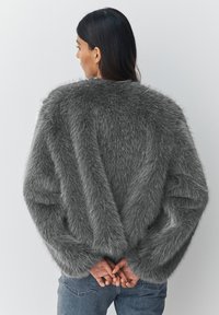 Woman with long dark hair wearing a plush gray faux fur jacket and blue jeans, standing with hands clasped behind back against white wall.