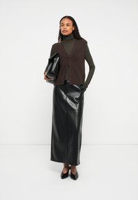 Kobieta w brązowej kamizelce bez rękawów, oliwkowym golfie, czarnej skórzanej spódnicy maxi i czarnych szpilkach, trzymająca dużą czarną torebkę kopertową.