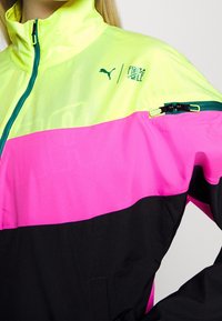 Colorful jacket with bright yellow, pink, and black panels. Features a high collar, half-zip closure, and side zip pocket. Made of lightweight fabric.