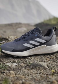 Chaussure de randonnée Adidas gris foncé et noir avec des bandes blanches sur un terrain rocheux et un arrière-plan de montagne flou.