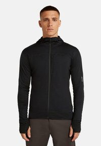 Hombre vestido con una chaqueta con capucha negra que se cierra con cremallera y tiene orificios para los pulgares, de pie frente a un fondo claro y liso, mirando hacia adelante.