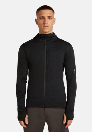 Man in een zwarte rits-hooded jacket met duim gaten, staand tegen een eenvoudige lichte achtergrond, met het gezicht naar voren.
