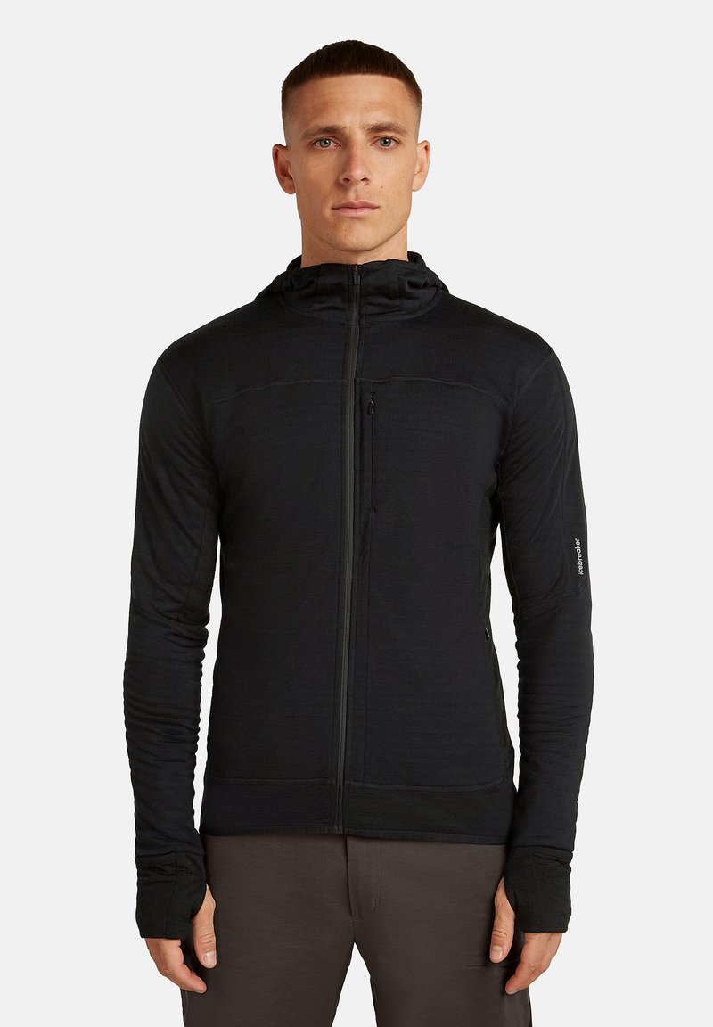 Hombre vestido con una chaqueta con capucha negra que se cierra con cremallera y tiene orificios para los pulgares, de pie frente a un fondo claro y liso, mirando hacia adelante.