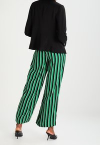 Svart blazer bärs över gröna och svarta randiga vida byxor. Byxorna har vertikala ränder i varierande bredd. Svarta klackskor.