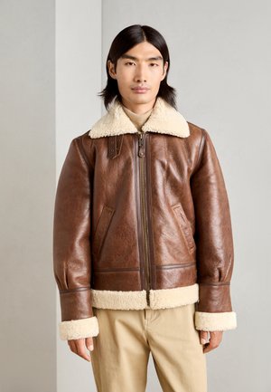 Veste en cuir marron avec un col et des poignets en shearling beige, fermeture éclair et deux poches latérales. Texture robuste et design ajusté.