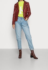 Rote Leder-Motorradjacke über einem leuchtend grünen gerippten Strickpullover, kombiniert mit hellblauen hochgeschnittenen Jeans und dunkelbraunen Stiefeletten.