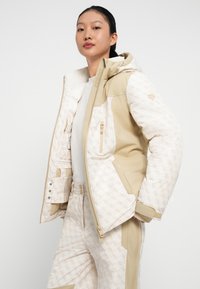 Veste de ski beige et crème avec un tissu matelassé et des accents de logo. Comprend une capuche, des poches zippées et une coupe ajustée, associée à un pantalon assorti.