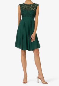 Smaragdgrön klänning med spetsoverdel, ärmlös design och flödande kjol. Åtföljd av klara högklackade sandaler.