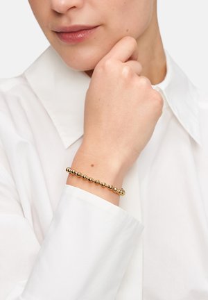 Goldene Perlenarmband an einem Handgelenk, mit abgerundeten Metallperlen und glatter Oberfläche, vor einem weißen Hemd-Hintergrund.
