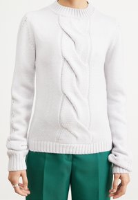 Svetle šedý pletený svetr s kabelovým vzorem uprostřed, žebrovaný kulatý výstřih a manžety, v kombinaci se zelenými kalhotami.