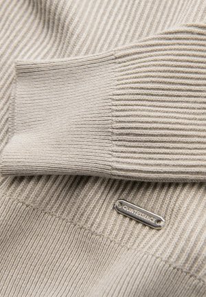 Beige geribbelde trui met een gladde textuur, voorzien van een zilverkleurig metalen label met de tekst "QUINTESSENCE." Close-up beeld legt de stikseldetails bloot.