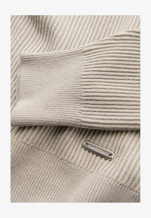 Beige geribbelde trui met een gladde textuur, voorzien van een zilverkleurig metalen label met de tekst "QUINTESSENCE." Close-up beeld legt de stikseldetails bloot.