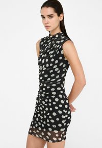 Schwarzes Kleid mit weißen Punkten, kurzen Ärmeln und gerafftem Design. Der Stoff scheint dehnbar zu sein, mit transparentem Saumdetail am unteren Rand.