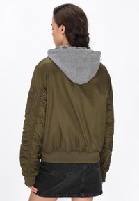 Giacca bomber verde oliva con cappuccio grigio, caratterizzata da polsini a costine e una finitura testurizzata. Include tasche laterali e una vestibilità rilassata.