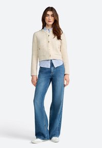 Cardigan corto color crema con due tasche frontali e bottoni dorati, indossato sopra una camicia a righe blu e abbinato a jeans a gamba larga.