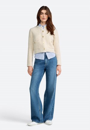 Cardigan corto color crema con due tasche frontali e bottoni dorati, indossato sopra una camicia a righe blu e abbinato a jeans a gamba larga.