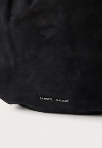 Sac en daim noir avec une texture lisse et un design minimaliste. Le nom de la marque "SAMØE SAMØE" est embossé en blanc sur le bas à l'avant.