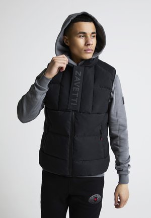 Młody mężczyzna w czarnej pikowanej kamizelce na szarym kapturze, poprawiający kaptur, patrzący lekko na bok na jednolitym tle.