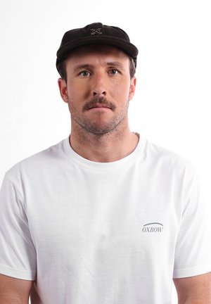 Homme avec des cheveux bruns courts et une moustache portant une casquette noire et un t-shirt blanc OXBOW, regardant droit devant lui sur fond blanc.