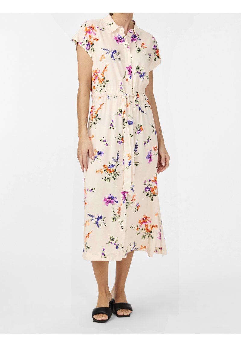 Femme portant une robe midi beige clair à imprimé floral coloré, manches courtes, boutonnée devant avec une ceinture à la taille, associée à des sandales noires.
