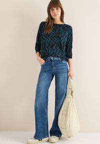 Zwart langärmelig top met een turkooise abstracte print, gecombineerd met lichtblauwe wijduitlopende jeans. Model draagt een crèmekleurige schoudertas. Casual schoeisel.