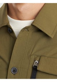 Zbliżenie na osobę w oliwkowej kurtce zapinanej na guziki z zamykanym na zamek kieszeniem, noszącą białą bluzkę z prążkowanej tkaniny.