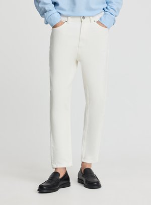 Persona che indossa pantaloni bianchi a gamba dritta, camicia a maniche lunghe azzurra, mocassini in pelle nera, mani nelle tasche, in piedi contro uno sfondo neutro.