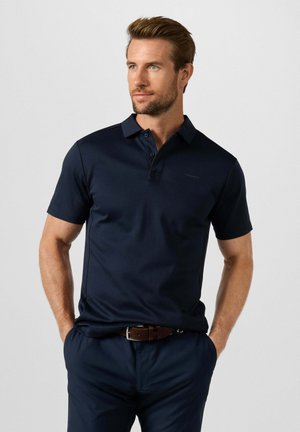 Man met kort haar en baard, draagt een donkerblauw poloshirt en bijpassende broek met een bruine riem, handen in de zakken, kijkt opzij.