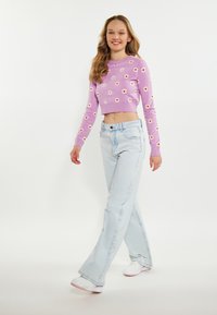 Pulover scurt de culoare violet deschis cu un model floral, purtat cu blugi evazați de culoare albastră deschis. Modelul poartă adidași albi. Fundal simplu.