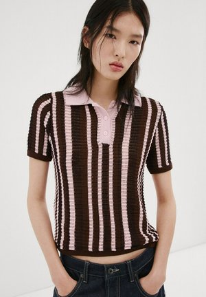 Mujer joven con una camiseta tipo polo de punto de manga corta con rayas verticales en marrón y rosa, con las manos en los bolsillos de unos vaqueros de mezclilla oscuros.