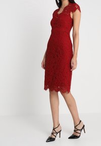 Femme portant une robe en dentelle rouge, courte à mi-cuisses et à manches courtes, associée à des chaussures à talons hauts noires à lacets, se tenant devant un fond blanc uni.