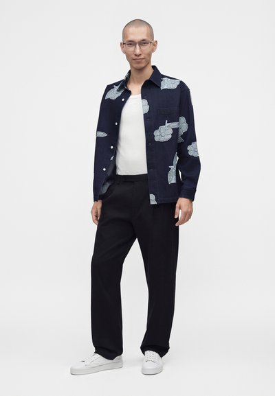 Homme portant des lunettes, veste bleu marine à motifs sur une chemise blanche, pantalon noir et baskets blanches, debout devant un fond uni.