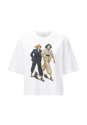 Deux femmes marchant avec confiance, portant des costumes élégants ceinturés — l'une en bleu marine foncé avec des lunettes de soleil, l'autre en beige — imprimées sur un t-shirt blanc.