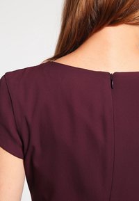 Robe bourgogne en tissu lisse et texturé, avec un décolleté large et un zip discret à l'arrière. Manches courtes.