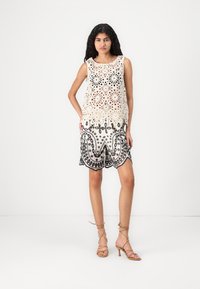Il top all'uncinetto presenta motivi floreali in beige, abbinato a pantaloni corti neri ricamati con bordi scallop. La modella indossa tacchi beige con cinturini.