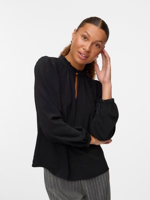 Black blouse with a ruffled neckline and keyhole detail, featuring long, puffed sleeves and a relaxed fit, styled with gray striped pants.
