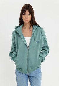 Svijetlosivi zip-up dukserica s opuštenim krojem, rebrastim manžetama i izvezenim logotipom. Nosena preko bijele majice i uparen sa svijetlim trapericama.
