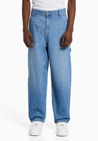 Osoba ubrana w jasnoniebieskie szerokie dżinsy i białe trampki, stojąca z rękami swobodnie wzdłuż ciała na jednolitym białym tle.