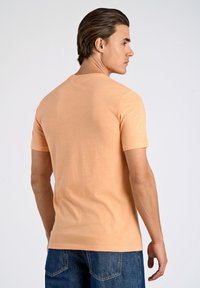Camiseta de manga corta en color durazno con cuello redondo, textura suave y diseño simple, mostrada desde la parte de atrás. Jeans azules incluidos.