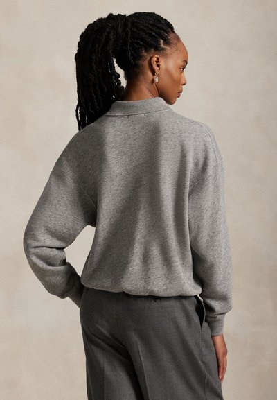 Woman with long braided hair tied back, wearing a gray sweater and gray pants, posed with one hand on her hip, facing sideways.