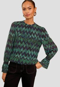 Bluza z dolgimi rokavi z zigzag vzorcem v zeleni in mornarsko modri barvi, z nabranim teksturo, okroglo ovratnico in volančki na koncu rokavov, okrašenimi z črnimi biseri.