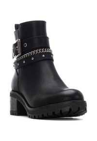 Botín negro de tobillo hecho de cuero sintético con un tacón grueso, que presenta una correa con hebilla, un acento de cadena plateada y detalles de tachuelas.