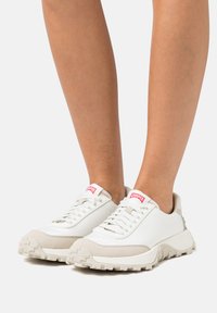 White sneakers with light beige accents; textured rubber sole, round toe, and fabric upper; features contrasting red logo on the tongue.