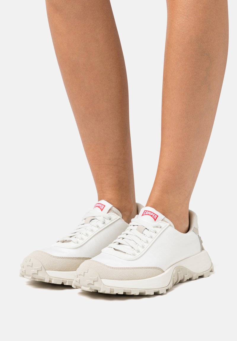 White sneakers with light beige accents; textured rubber sole, round toe, and fabric upper; features contrasting red logo on the tongue.