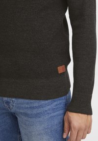 Pull gris foncé texturé avec un ourlet et des poignets côtelés, doté d'une étiquette en cuir marron sur le côté inférieur, porté avec un jean bleu.