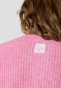 Ružový pletený sveter s rebrovanou textúrou, s štvorcovou visačkou označenou "LA MAISON AMV" v ružovej farbe na bielom pozadí.