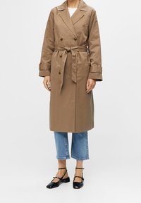 Mujer vestida con un trench beige de longitud a la rodilla, atado a la cintura, jeans cortos azules y bailarinas negras con tira en el tobillo, de pie contra un fondo blanco.