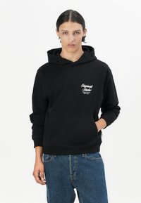 Black hoodie with a kangaroo pocket, featuring white script logo on the left chest. Fabric is soft, with ribbed cuffs and hem. Paired with blue jeans.